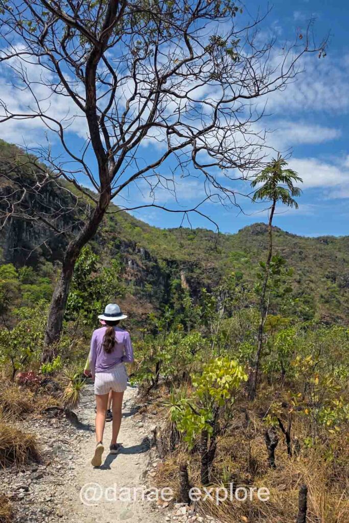 Moça com shorts, camiseta e chapéu caminhado por trilha plana em meio a vegetação do cerrado, na trilha amarela do Parque Nacional da Chapada dos Veadeiros