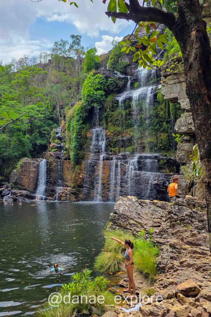Mulher de biquíni aponta para uma pessoa nadando em um poço de água escura, na Cachoeira Almécegas I, na Chapada dos Veadeiros.