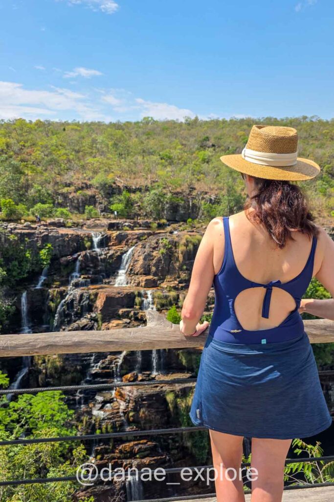 Eu, com chapéu e maiô azul de costas para câmera, observo de um mirante as diversas quedas d’água de uma grande cachoeira - Cachoeira Almecégas 1, cercada por vegetação do cerrado.
