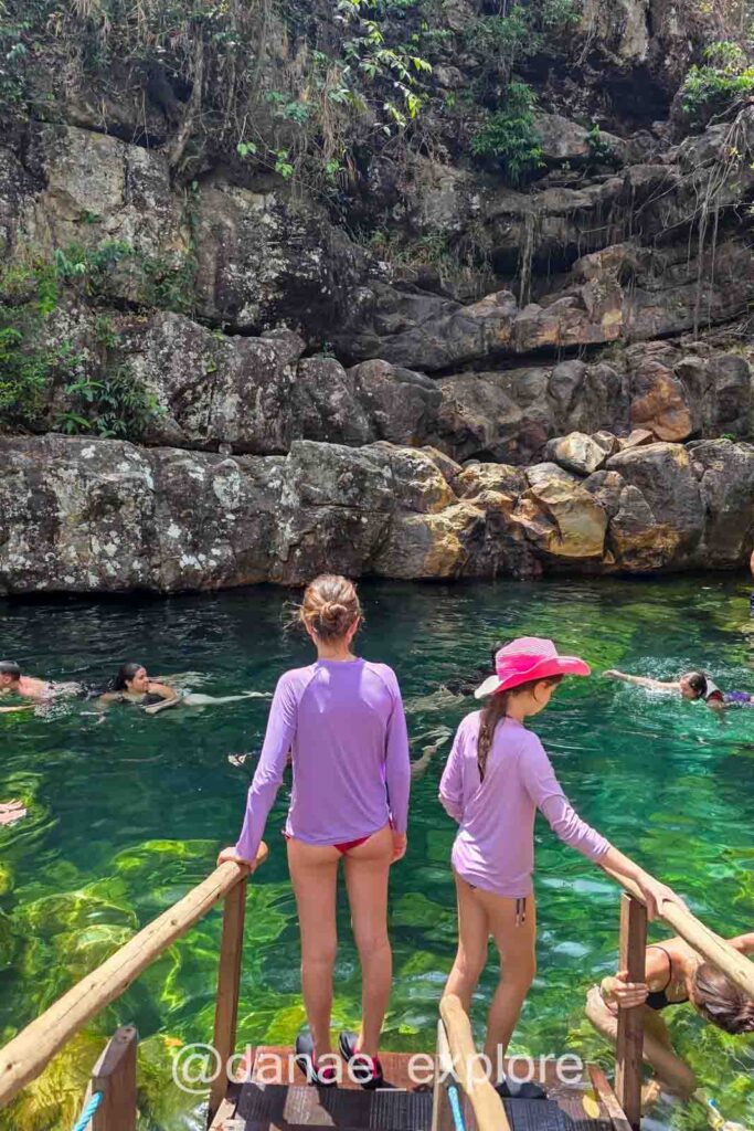 Poço do Xamã, na Cachoerias Loquinhas. Duas meninas em passarela de acesso ao poço de águas cristalinas esverdeadas, em dia de sol. Há outras pessoas nadando no poço.