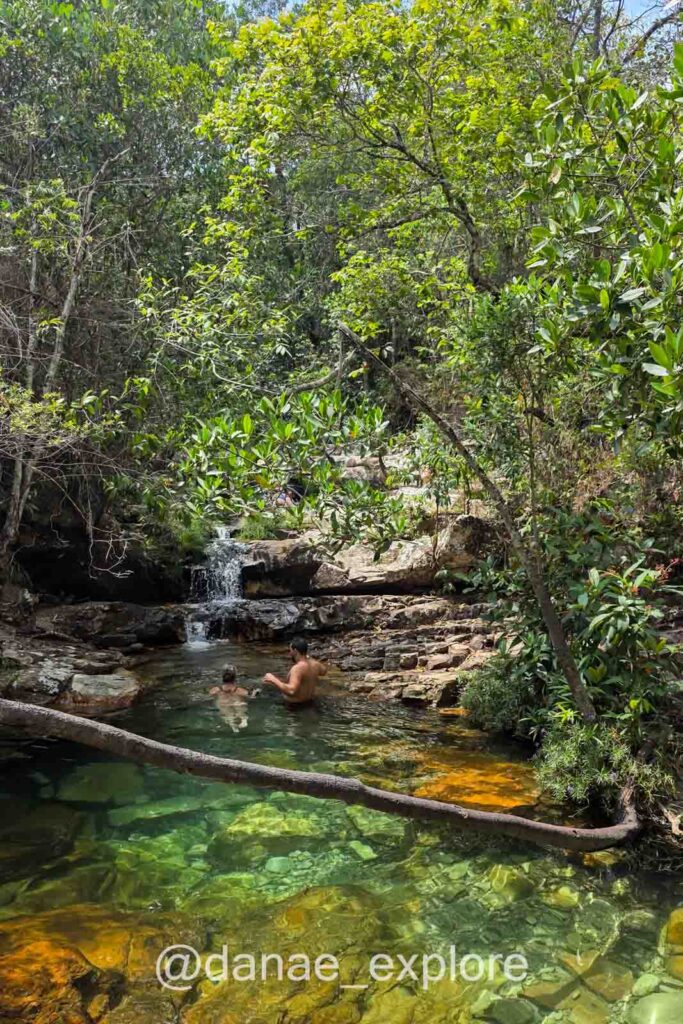 Duas pessoas relaxam em um pequeno poço natural de águas transparentes com fundo dourado, em meio à vegetação nativa e uma queda d’água suave, nas Cachoeiras Loquinhas, na Chapada dos Veadeiros.