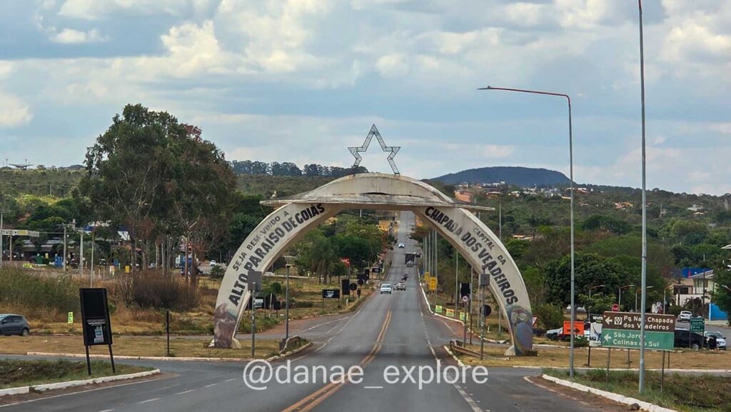 Pórtico de entrada da cidade de Alto Paraíso de Goiás com a frase “Seja bem-vindo à capital da Chapada dos Veadeiros” em destaque, sobre uma estrada rodeada por vegetação em dia de sol com nuvens