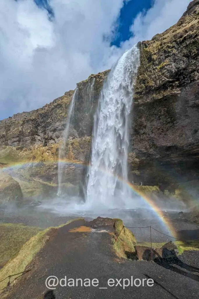 Seljalandsfoss waterfall with a rainbow formed by the mist of the water, trail passing behind the falls.
