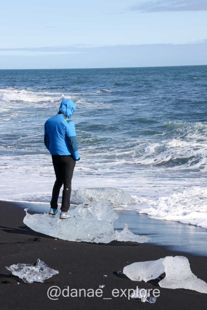 pessoa em casaco azul e calça preta em cima de gelo a beira do mar, com ondas fortes a se aproximar, na Diamond Beach