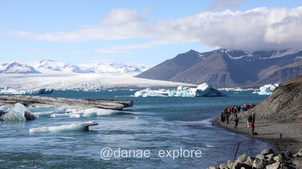 Lagoa Jökulsárlón em dia claro, com céu azul. Há diversos icebergs na lagoa e alguns turistas nas margens. Ao fundo, o Glaciar Vatnajökull.