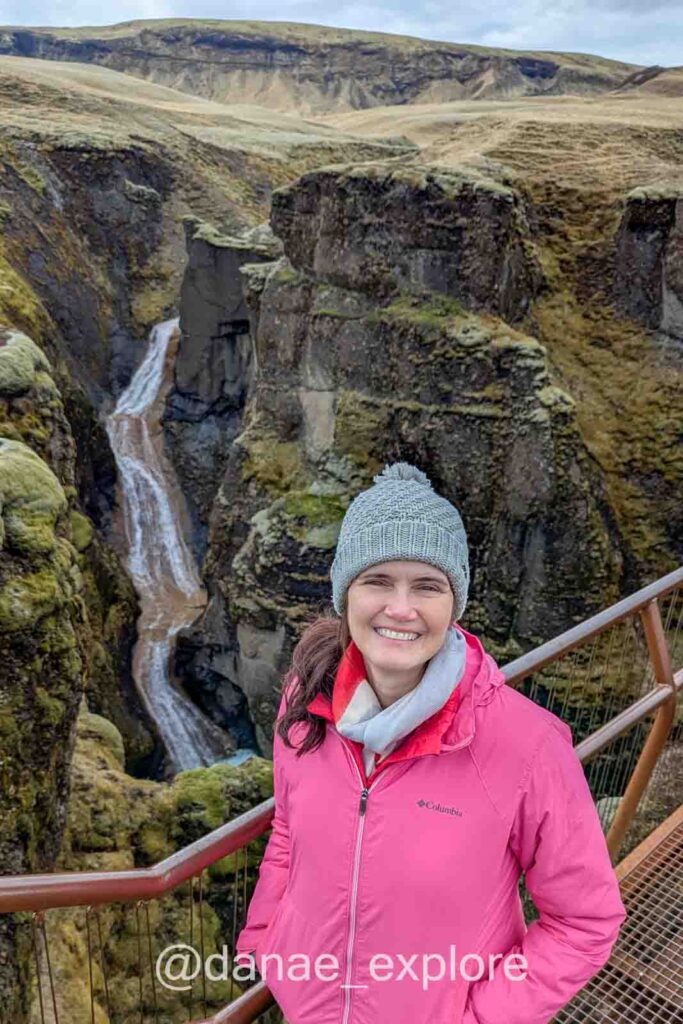 Eu estou sorrindo para câmera no mirante para o cânion Fjaðrárgljúfur, com cachoeira Mogafoss ao fundo e paisagem verde musgosa