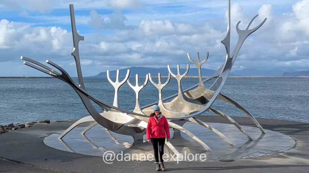 Me standing in front of the Sun Voyager sculpture by the sea in Reykjavík, with mountains in the background. It's a cold and cloudy day, but some sun is shining on the sculpture.