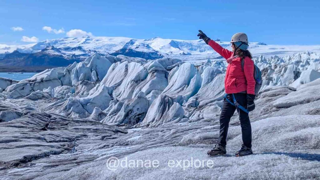 Eu, com casaco vermelho, calça preta e equipamento de caminhada no gelo, na geleira Vatnajökull, apontando em direção ao Lago Jökulsárlón, que aparece ao fundo