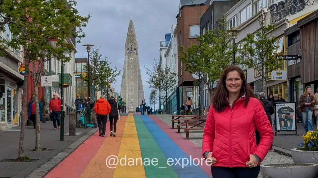 I'm wearing a red winter sweater, posing for a photo on Rainbow Street, with Hallgrímskirkja Church in the background, in Reykjavík. There are other tourists walking along the street.