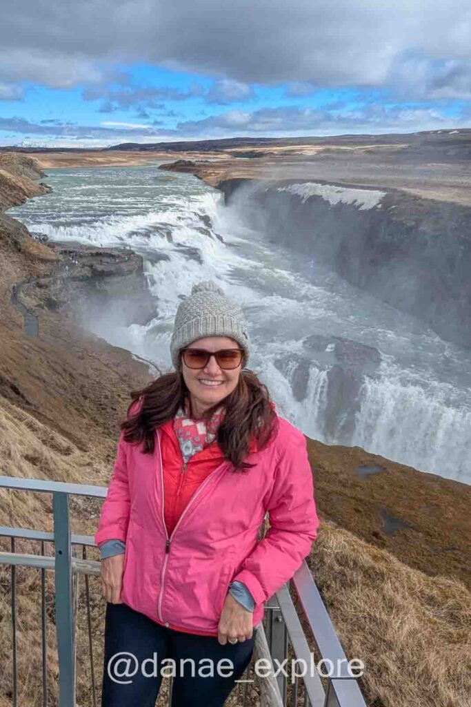 Eu, com blusa rosa, gorro e óculos de sol, sorrio para câmera em frente a Gullfoss, parte essencial do que sugiro fazer na Islândia