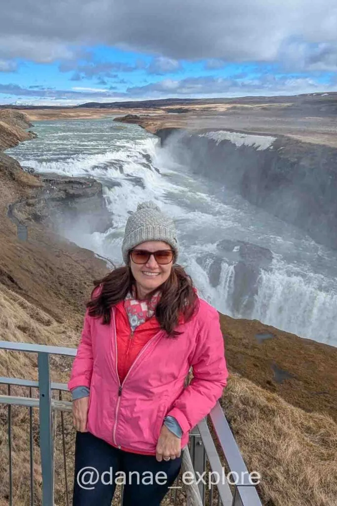 I, wearing a pink blouse, beanie, and sunglasses, smile at the camera in front of Gullfoss, an essential part of what I suggest doing in Iceland.