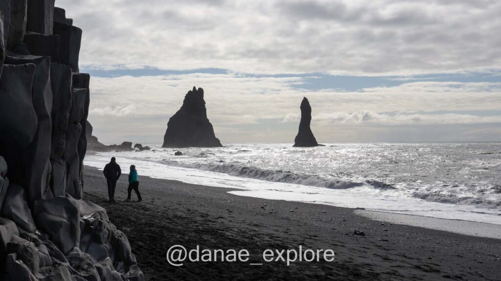 Praia de areia preta de Reynisfjara com colunas de basalto e rochedos no mar, sul da Islândia. Está nublado e a imagem parece em preto-e-branco, mas há um rasgo de céu azul