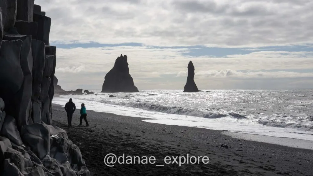 Black sand beach of Reynisfjara with basalt columns and rocks in the sea, southern Iceland. It's cloudy and the image appears in black and white, but there's a patch of blue sky.