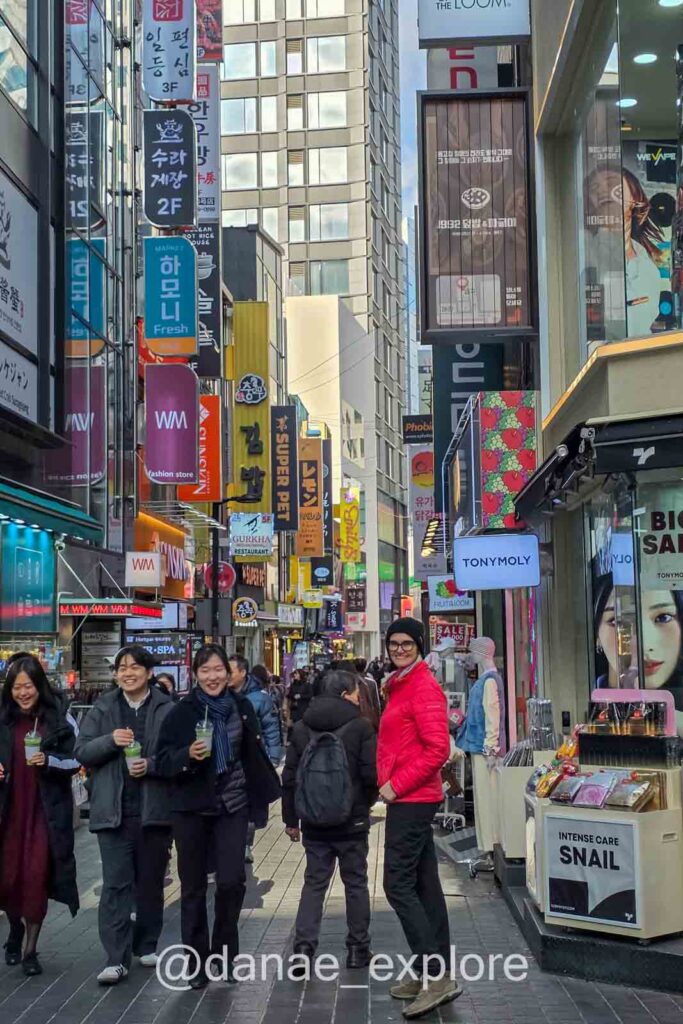 Eu, com meu casaco vermelho, passendo pela rua de Myeongdong em Seul com lojas, letreiros coreanos e movimento intenso de pedestres