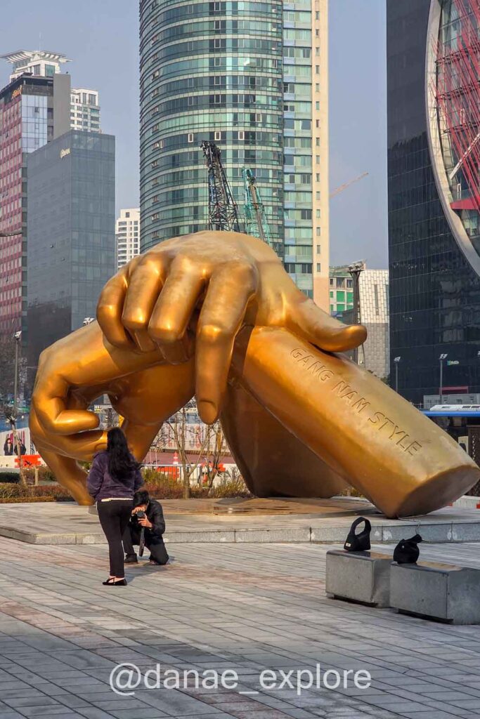 Pessoas tiram fotos em escultura Gangnam Style em Seul com mãos douradas gigantes em praça urbana cercada por prédios