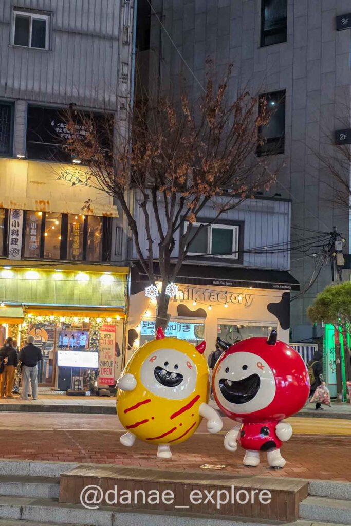 Esculturas coloridas em rua comercial de Hongdae à noite com lojas iluminadas e clima urbano em Seul