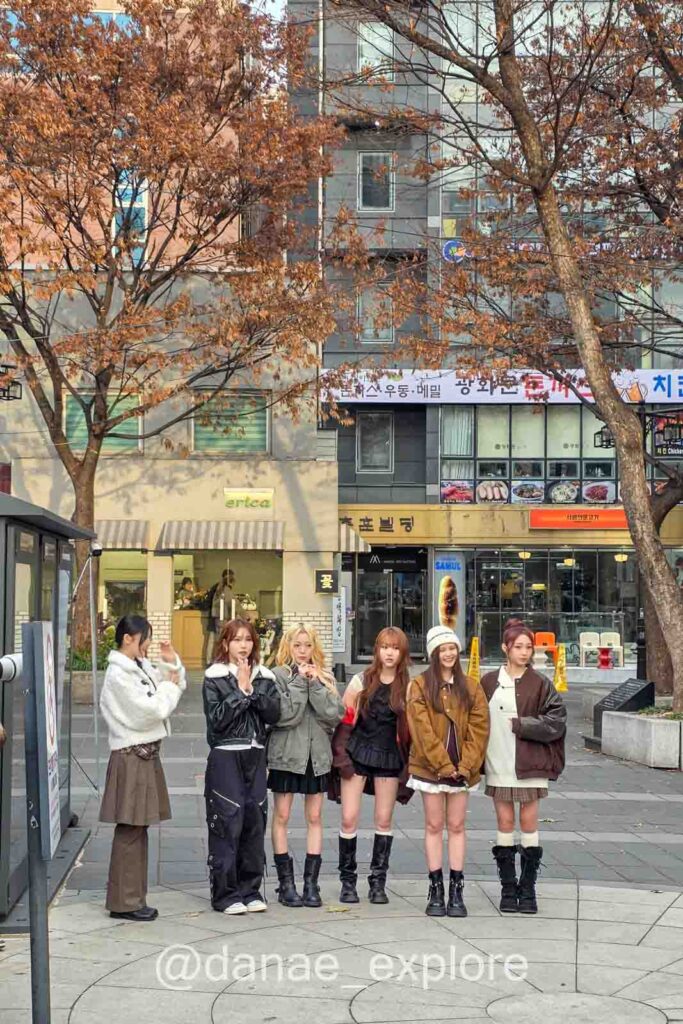 Jovens cantoras de grupo de K-pop se preparam para apresentação em rua de Seul com lojas, cafés e árvores de outono ao redor