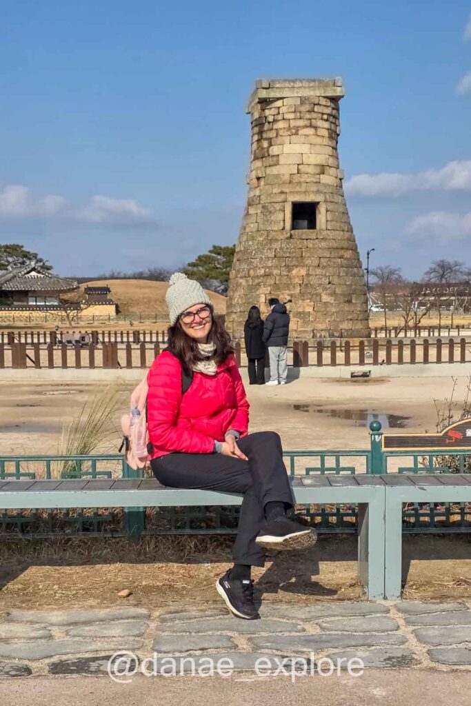 Eu, com blusa vermelha e gorro, sentada em banco a frente do Observatório Cheomseongdae em Gyeongju com torre histórica de pedra em área arqueológica aberta