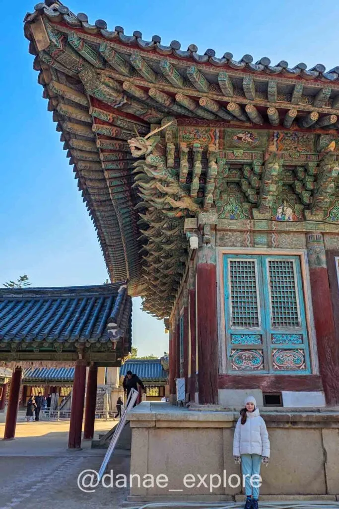 Criança posa para foto na lateral do templo Bulguksa, em Gyeongju. É possível ver detalhe da pintura dancheong colorida e a arquitetura tradicional