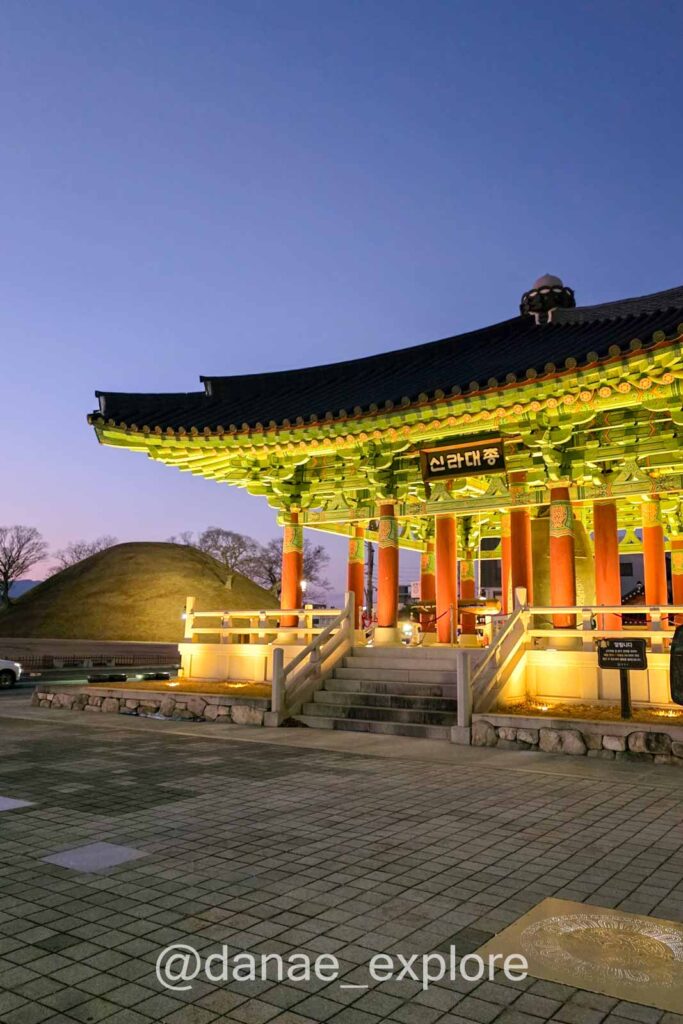 Pavilhão iluminado em Gyeongju ao anoitecer com arquitetura tradicional coreana em destaque. Ao fundo, colina de uma tumba do reino de Silla.