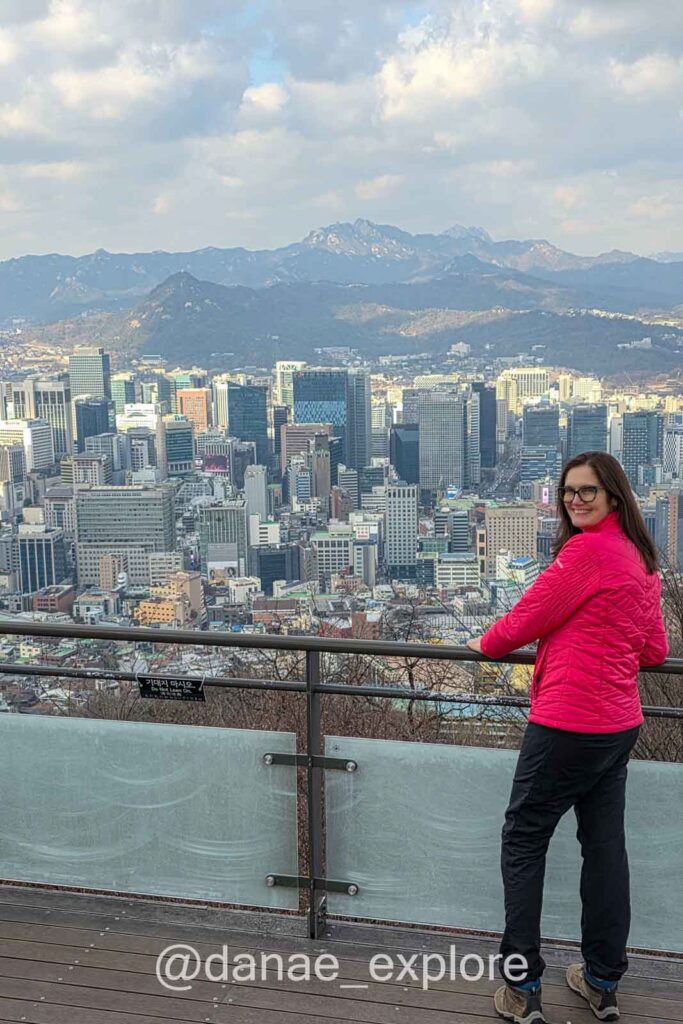Eu, em jaqueta vermelha e calça preta, observo prédios modernos de Seoul no fim da tarde, do parque da N-Seoul Tower. Ao fundo é possível ver montanhas