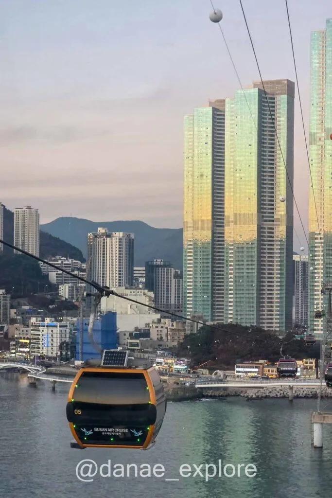 Fim de tarde no Songdo Cable Car (Busan Air Cruise), sobre o mar com prédios modernos e montanhas ao fundo