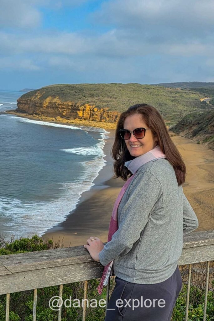 Moça de óculos de sol sorri para câmera, ao fundo a praia Torquay, uma das primeiras paradas da Great Ocean Road
