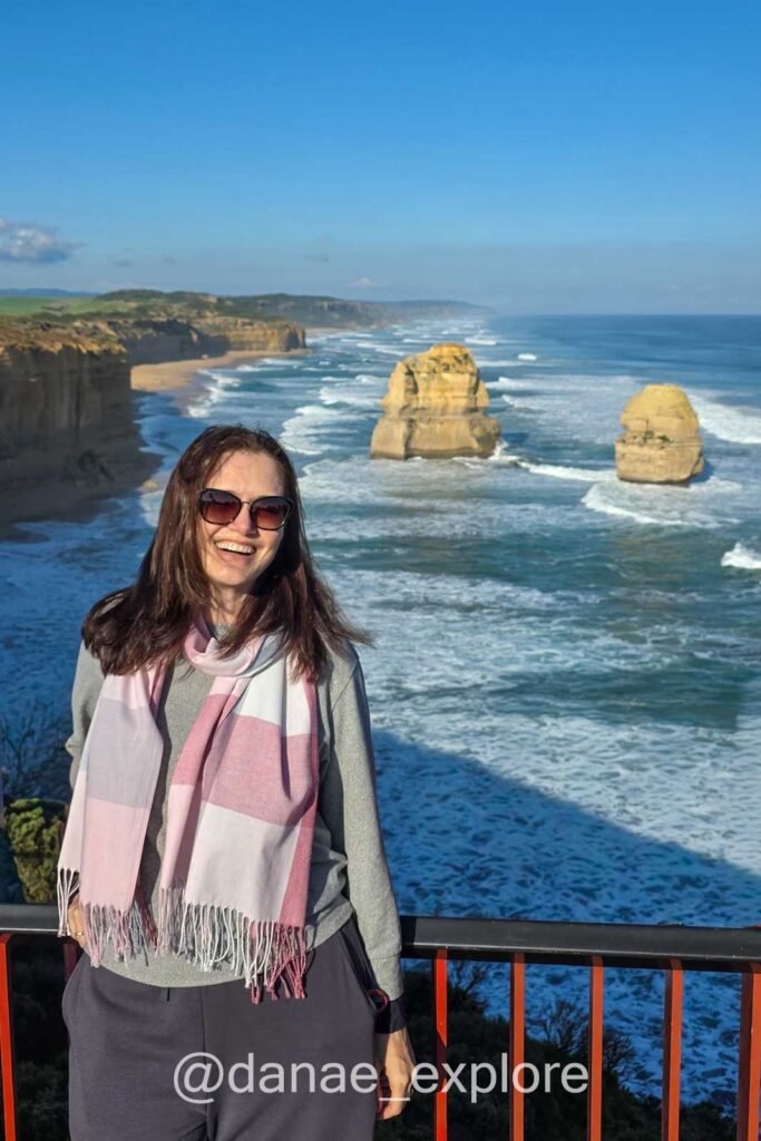 Moça de óculos de sol sorri para câmera, ao fundo praia com ondas e algumas das formações rochosas dos Doze Apóstolos, na Great Ocean Road