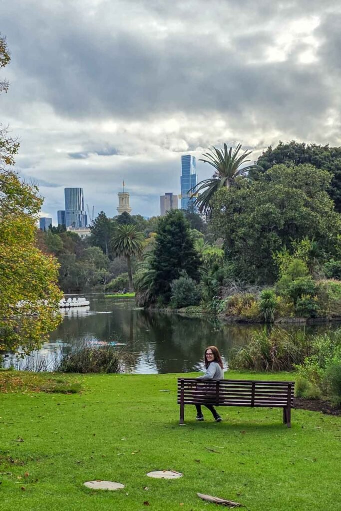 Moça olha para trás, em direção à câmera, no Royal Botanic Gardens em Melbourne, ao fundo se veem alguns arranha-céus, em dia nublado.
