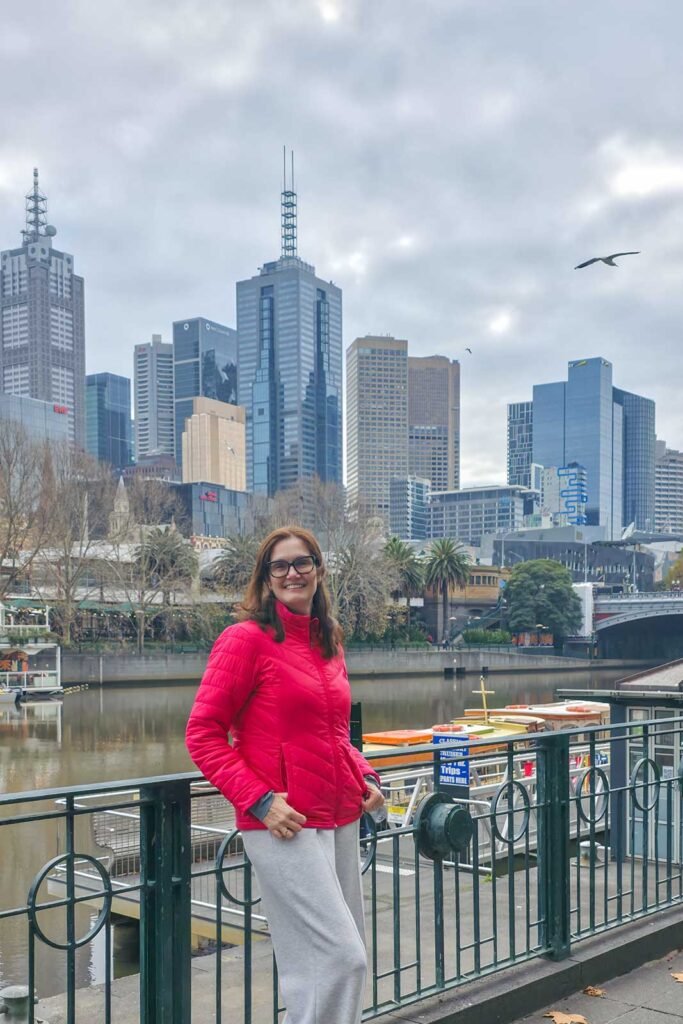 Moça com jaqueta vermelha as margens do Rio Yarra, com o skyline de Melbourne ao fundo em dia nublado