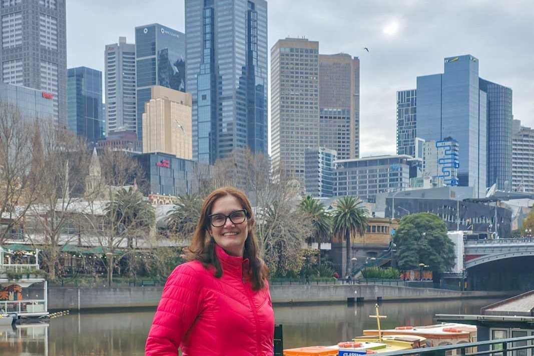 Eu, com jaqueta vermelha, as margens do Rio Yarra com o skyline de Melbourne ao fundo, em dia nublado