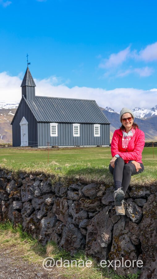 Eu, com casaco vermelho, calça preta e gorro, sentada na grama em frente a Igreja Budakirkja, na península Snæfellsnes na Islândia