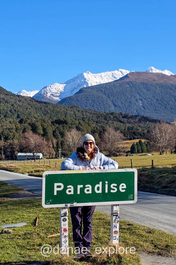 Dânae atrás da placa de Paradise, com campos e montanhas nevadas ao fundo, que visitamos como bate-volta de Queenstown