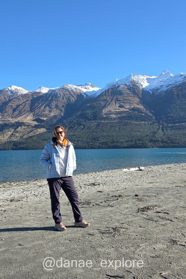 Dânae na margem do Lago Wakatipu, com montanhas nevadas do outro lado da água.