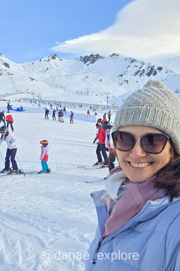 Dânae sorri na área de esqui de The Remarkables, com esquiadores e montanhas cobertas de neve ao fundo.