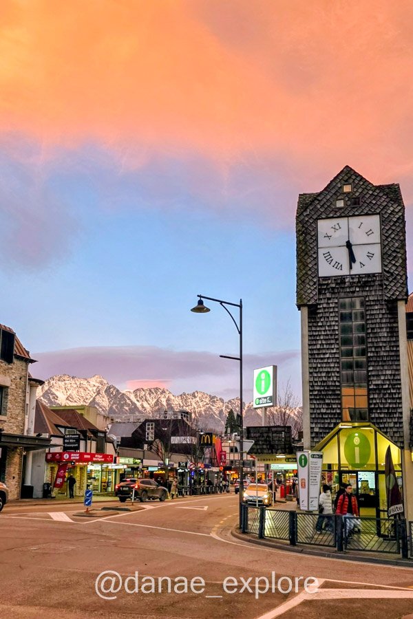 Torre do relógio no centro de Queenstown, com montanhas nevadas e céu alaranjado ao entardecer.