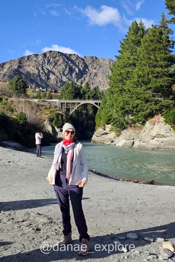 Eu na margem do rio Shotover, prestes a embarcar no Shotover Jet, com uma ponte em arco e montanhas ao fundo.