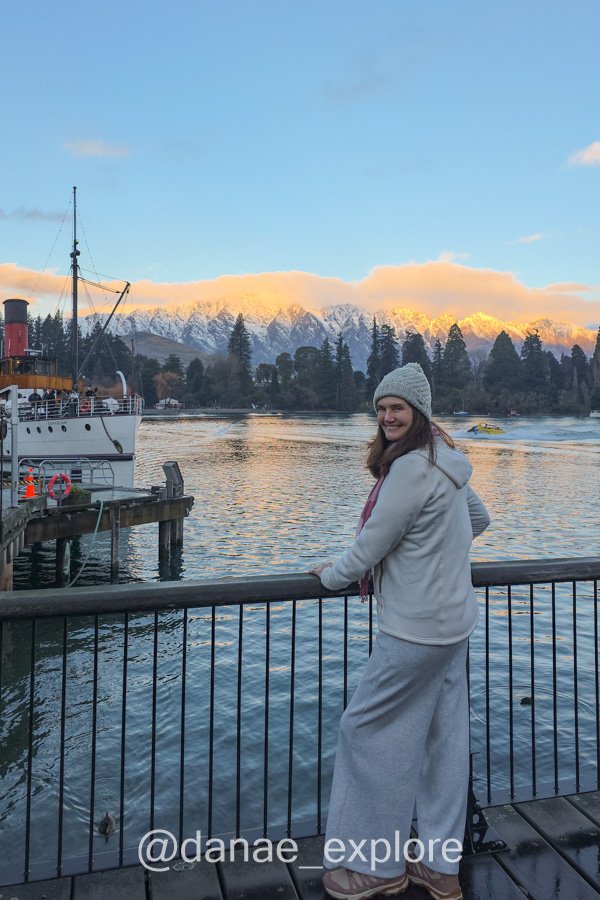 Em Queenstown, estou junto ao Lago Wakatipu, com o barco a vapor TSS Earnslaw e montanhas nevadas ao fundo, num fim de tarde de inverno