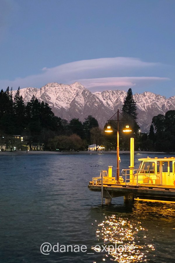 Barco iluminado no Lago Wakatipu ao anoitecer, com montanhas nevadas ao fundo.