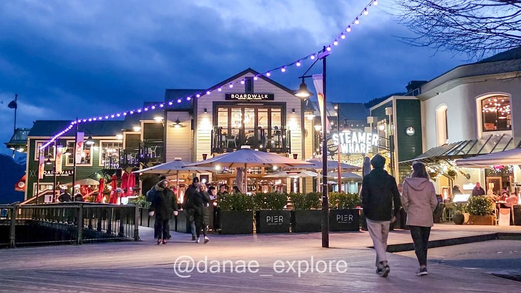 Pessoas caminham pelo Steamer Wharf, no centro de Queenstown, sob luzes acesas ao anoitecer.