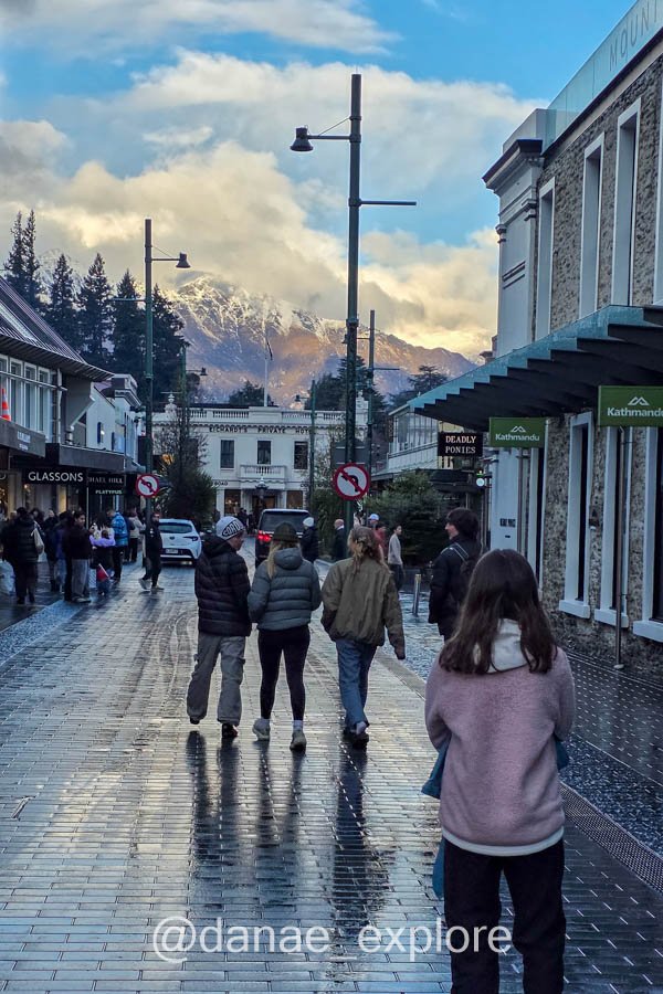 Pessoas caminham por uma rua molhada no centro de Queenstown, com montanhas ao fundo.