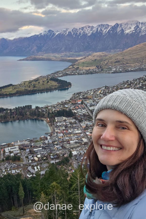 Eu, de gorro e casaco, no mirante do Bob's Peak, com vista de Queenstown, com o Lago Wakatipu e montanhas nevadas ao fundo.